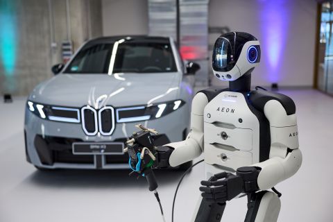 Photo of white humanoid robot holding a tool in front of a shiny new grey car