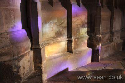 purple, yellow and green light shining through a stained glass window onto the stonework of Gloucester Cathedral